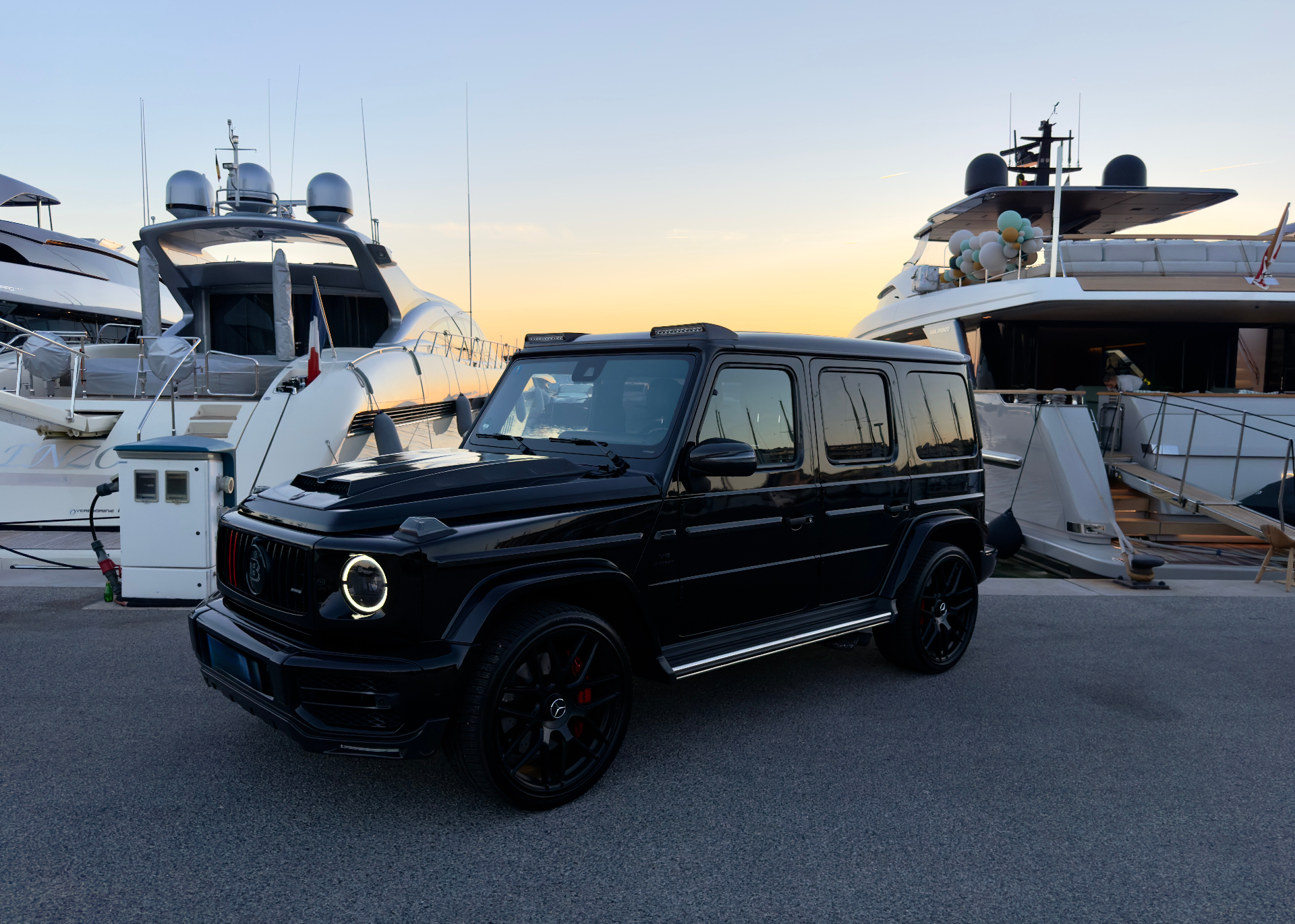 Mercedes-AMG G63 Brabus alongside superyachts at a French Riviera marina at sunset